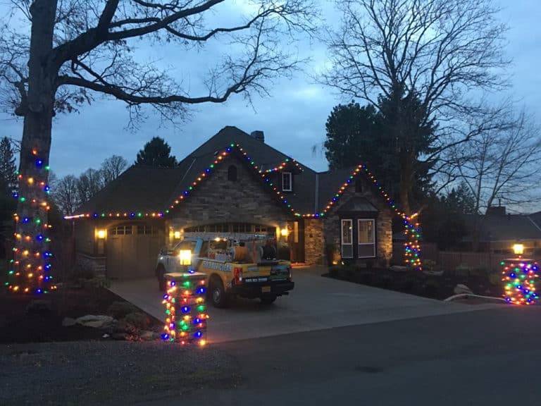 Home with multi-colored holiday lights along the roofline and wrapped trees at dusk